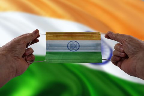 Das Bild zeigt zwei Hände, die eine OP-Maske in den indischen Nationalfarben halten. Im Hintergrund weht die indische Flagge.