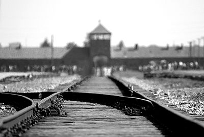 Das Bild zeigt das Torhaus des Konzentrationslager Auschwitz Birkenau und darauf zulaufende Eisenbahnschienen.