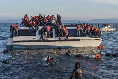 das Bild zeigt ein kleines Boot, das völlig mit Menschen mit und ohne Schwimmwesten überfüllt ist. Helfer eilen vom Ufer her zu den gestrandeten.