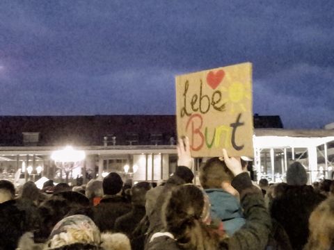 Das Bild zeigt ein Pappschild mit der Aufschrift "Lebe bunt!" in verschiedenfarbigen Buchstaben geschrieben. Das Schild wird inmitten einer Menschenmenge hochgehalten.